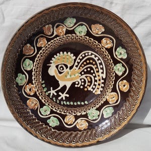 May include: A decorative ceramic plate with a brown background and a detailed rooster design in the center. The plate features a border of floral and geometric patterns in shades of brown, white, green, and orange.