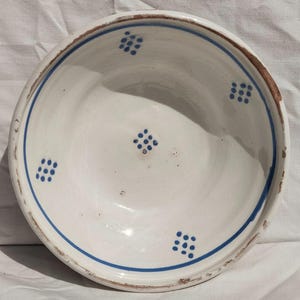 May include: A round ceramic bowl with a white glazed interior and exterior. The rim features a thin blue painted line, and the interior is decorated with five small blue dot clusters arranged in a diamond pattern. The edges show signs of wear.