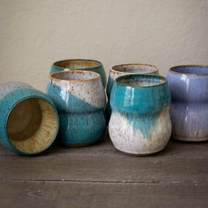 May include: A collection of handmade ceramic tumblers in various shades of blue, turquoise, and white. The tumblers have a unique, rounded shape and a glossy finish. Some have a two-tone design, while others are solid colors.
