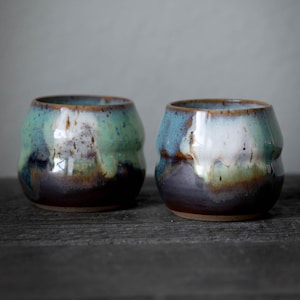 K&ouml;nnte beinhalten: Zwei handgefertigte Keramikbecher mit einer einzigartigen Glasur. Die Becher haben eine abgerundete Form und eine strukturierte Oberfl&auml;che. Die Glasur weist eine Mischung aus Farben auf, darunter T&uuml;rkis, Braun und Wei&szlig;, wodurch ein Marmoreffekt entsteht. Die Becher stehen auf einer dunklen Holzoberfl&auml;che.