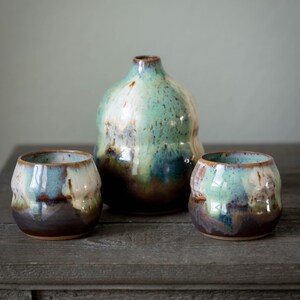 Könnte beinhalten: Ein Set aus drei handgefertigten Keramikteilen mit glänzendem, strukturiertem Finish. Das Set beinhaltet eine bauchige Vase und zwei kleine, abgerundete Tassen. Die Stücke weisen eine Mischung aus Türkis, Braun und Creme auf.
