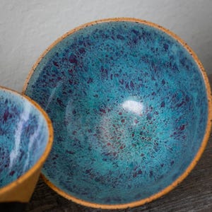 May include: Two ceramic bowls with a speckled turquoise glaze. The bowls have a rounded shape with a light brown rim. The interior of the bowls have a glossy finish with dark purple speckles. The bowls are set on a dark wooden surface.