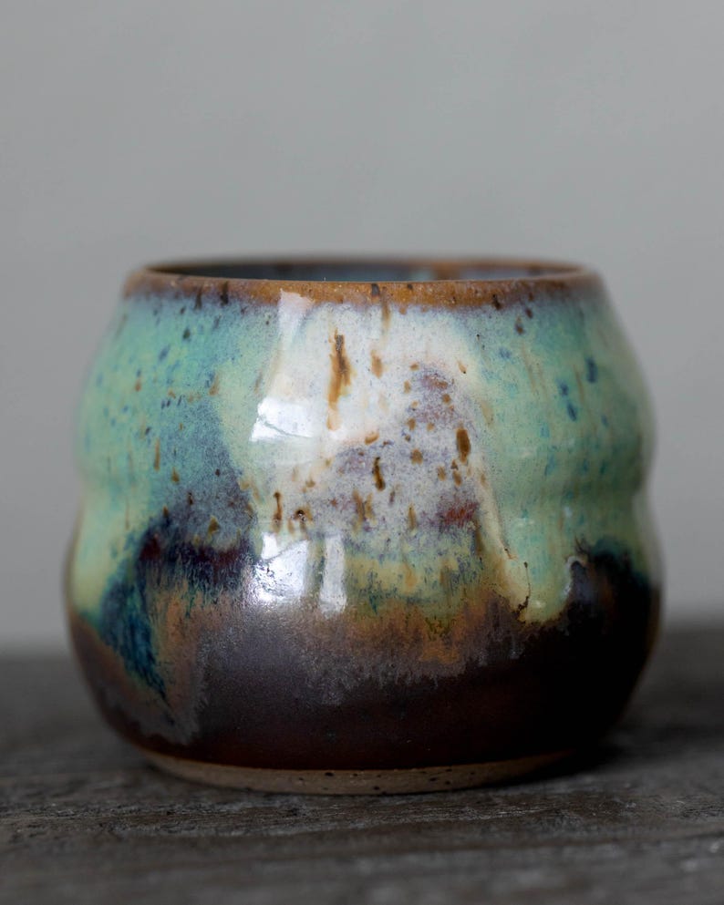 K&ouml;nnte beinhalten: Eine handgefertigte Keramiktasse mit abgerundeter Form. Die Tasse weist eine Mischung aus Farben auf, darunter Braun, T&uuml;rkis und Wei&szlig;, mit gl&auml;nzendem Finish. Der Rand ist dunkler braun, und das Gesamtdesign ist organisch und strukturiert.