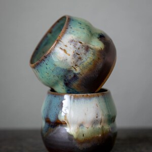 K&ouml;nnte beinhalten: Zwei handgefertigte Keramikbecher, die &uuml;bereinander gestapelt sind. Die Becher haben eine einzigartige Form und sind mit einer Mischung aus Farben glasiert, darunter T&uuml;rkis, Braun und Wei&szlig;. Die Becher werden wahrscheinlich zum Trinken von Getr&auml;nken verwendet.