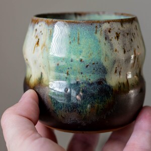 K&ouml;nnte beinhalten: Eine handgefertigte Keramiktasse mit einer einzigartigen Glasur. Die Tasse weist eine Mischung aus Braun-, Gr&uuml;n- und Cremet&ouml;nen auf. Die Au&szlig;enseite hat ein strukturiertes, organisches Aussehen. Die Tasse wird in einer Hand gehalten.