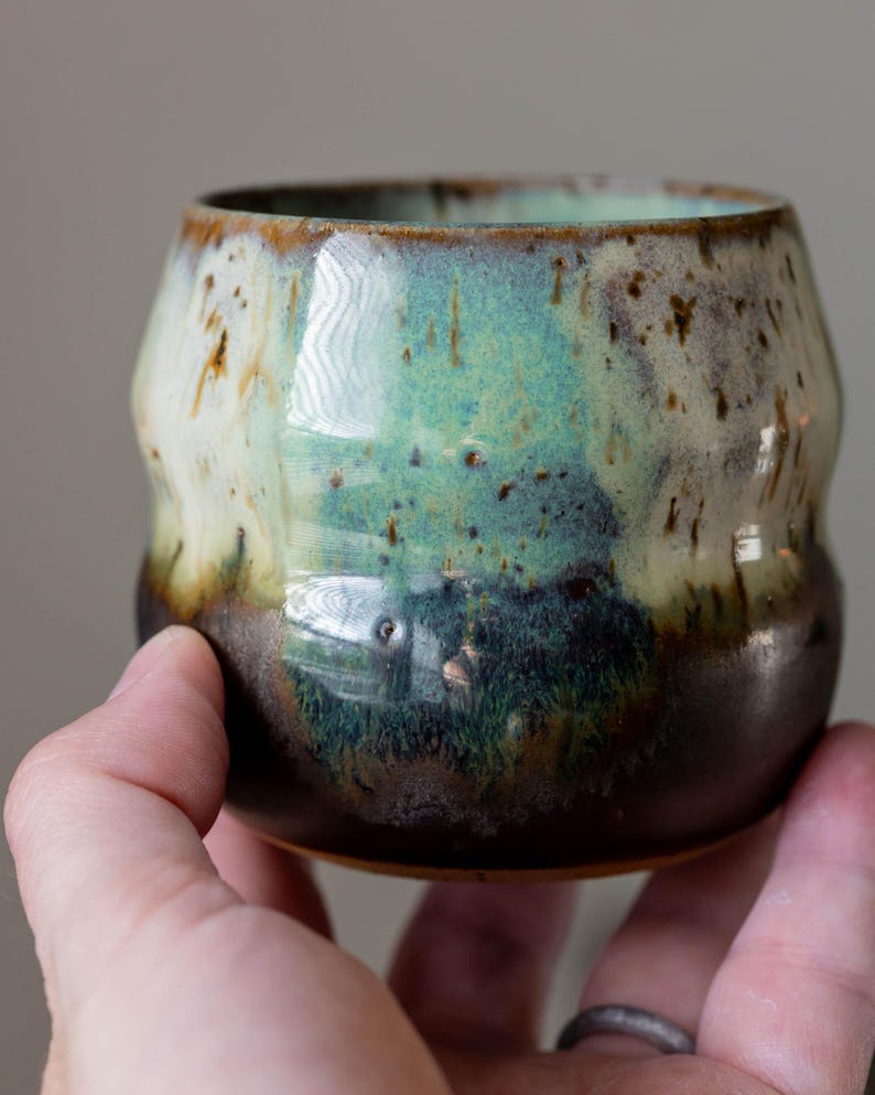 K&ouml;nnte beinhalten: Eine handgefertigte Keramiktasse mit einer einzigartigen Glasur. Die Tasse weist eine Mischung aus Braun-, Gr&uuml;n- und Cremet&ouml;nen auf. Die Au&szlig;enseite hat ein strukturiertes, organisches Aussehen. Die Tasse wird in einer Hand gehalten.