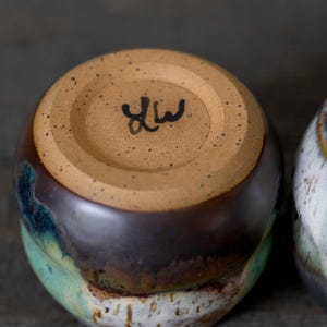 K&ouml;nnte beinhalten: Eine handgefertigte Keramiktasse mit brauner, blaugr&uuml;ner und wei&szlig;er Glasur. Der Boden der Tasse ist unglasiert und tr&auml;gt eine gestempelte Signatur. Die Tasse hat eine abgerundete Form und dient wahrscheinlich zum Trinken von Getr&auml;nken.