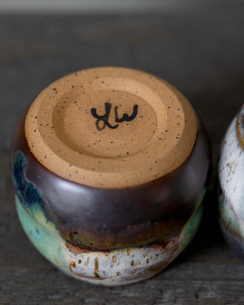 K&ouml;nnte beinhalten: Eine handgefertigte Keramiktasse mit brauner, blaugr&uuml;ner und wei&szlig;er Glasur. Der Boden der Tasse ist unglasiert und tr&auml;gt eine gestempelte Signatur. Die Tasse hat eine abgerundete Form und dient wahrscheinlich zum Trinken von Getr&auml;nken.