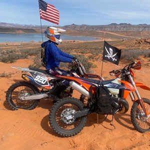 May include: Two orange and black dirt bikes parked on a red sand dune with a blue sky and mountains in the background. The dirt bikes have a black backpack on one and a black and white pirate flag on the other. The rider is wearing a blue jacket and a white helmet with a 100% logo.