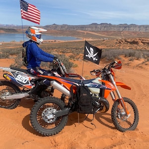 May include: Two orange dirt bikes parked on a red sand dune with a blue sky and mountains in the background. The dirt bikes have black seats and black tires. A black pirate flag with a skull and crossbones is attached to the handlebars of one of the bikes. An American flag is attached to the handlebars of the other bike.