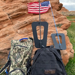 May include: Two camouflage backpacks, one black backpack, and two black flag poles with blue and black flags attached. The flags are hanging from the poles. The scene is set against a red rock background.