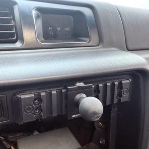 May include: A black dashboard with a digital clock displaying "H M :00". A black mounting bracket with a textured ball-shaped knob is attached to the dashboard. The dashboard also has air vents and control buttons.