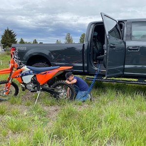 May include: A young person kneeling and using an air hose to inflate the tire of an orange and black dirt bike. The bike is parked next to a gray pickup truck with the door open.
