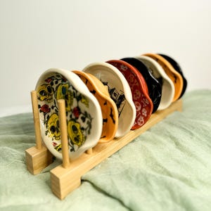 May include: A set of seven small, round ceramic dishes with Halloween-themed designs, including skulls, bats, and haunted houses. The dishes are displayed on a natural wood rack, showcasing a variety of colors and patterns.