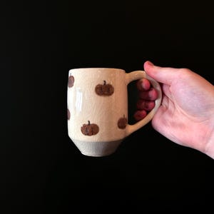 May include: A white ceramic mug with a brown pumpkin design. The mug has a handle and a slightly rounded bottom.