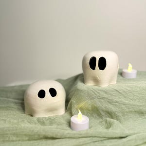 May include: Two off-white ceramic ghost figurines with black eye cutouts sit on a sage green cloth. Battery-operated tea lights are placed around the ghosts, creating a spooky Halloween theme.