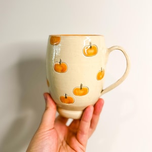 May include: Cream-colored ceramic mug decorated with orange pumpkin illustrations. The mug has a rounded body, a handle, and a slightly flared base. The pumpkins are outlined in black and have orange stems. The interior and rim of the mug are also orange.