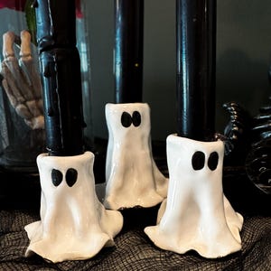 May include: Three white ceramic ghost candle holders with black eyes, each holding a black candle. The holders are on a black mesh surface, creating a spooky Halloween theme.