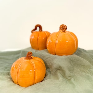 May include: Three ceramic pumpkins in varying sizes, each with a glossy orange finish and a brown stem. The pumpkins are arranged on a sage green fabric, creating a seasonal autumn theme. The pumpkins have vertical ridges.