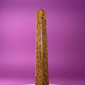 May include: A tall, rectangular obelisk with a polished surface. The stone features a swirling pattern of brown and gold, creating a marbled effect. The obelisk stands against a solid purple background.