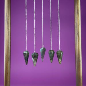 May include: A wooden pendulum stand with five gemstone pendulums hanging from silver chains. The pendulums are dark green and faceted. The stand has a dark brown top and light brown posts, set against a purple background.