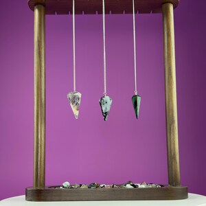 May include: Three gemstone pendulums hanging from a wooden display stand. The pendulums are different colors: clear, teal, and green. The stand is brown with a purple background. A small collection of stones is at the base of the stand.
