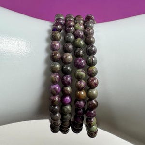May include: A set of five beaded bracelets. Each bracelet is made of small, round beads in shades of purple, green, and gray. The bracelets are displayed on a white form against a pink background. The beads appear to be approximately 0.3 inches in diameter.