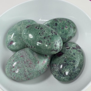May include: Five polished oval-shaped stones. The stones are a mottled green color with small red inclusions. The stones are smooth and shiny, and they are arranged in a white bowl. The stones are a natural product.