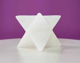 May include: A white, geometric star-shaped sculpture with a faceted design. The sculpture is made of a solid, opaque material and sits on a white surface against a purple background. The star has six points and a three-dimensional appearance.