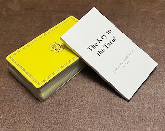 May include: A yellow tarot deck with a black geometric design and a white book titled "The Key to the Tarot" with the subtitle "Based on The Pictorial Key to the Tarot". The items are on a brown surface.