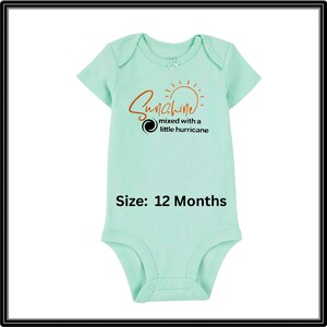Może przedstawiać: Jasnozielone body dla niemowląt z kr&oacute;tkim rękawem. Z przodu napis "Sunshine mixed with a little hurricane" pomarańczową czcionką i grafika słońca. Rozmiar: 12 miesięcy.