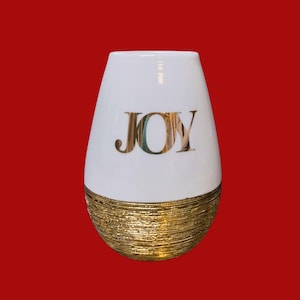 May include: A white ceramic vase with a tapered, rounded shape. The upper portion is smooth white with the word JOY printed in large, reflective gold capital letters. The lower portion features a textured, horizontal gold metallic finish.