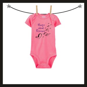 Pode incluir: Um body rosa para beb&eacute; com as palavras "Hugs and Kisses" e desenhos de cora&ccedil;&otilde;es estampados na frente. O body est&aacute; pendurado num varal com molas de madeira. O body &eacute; de manga curta e tem fecho de press&atilde;o.