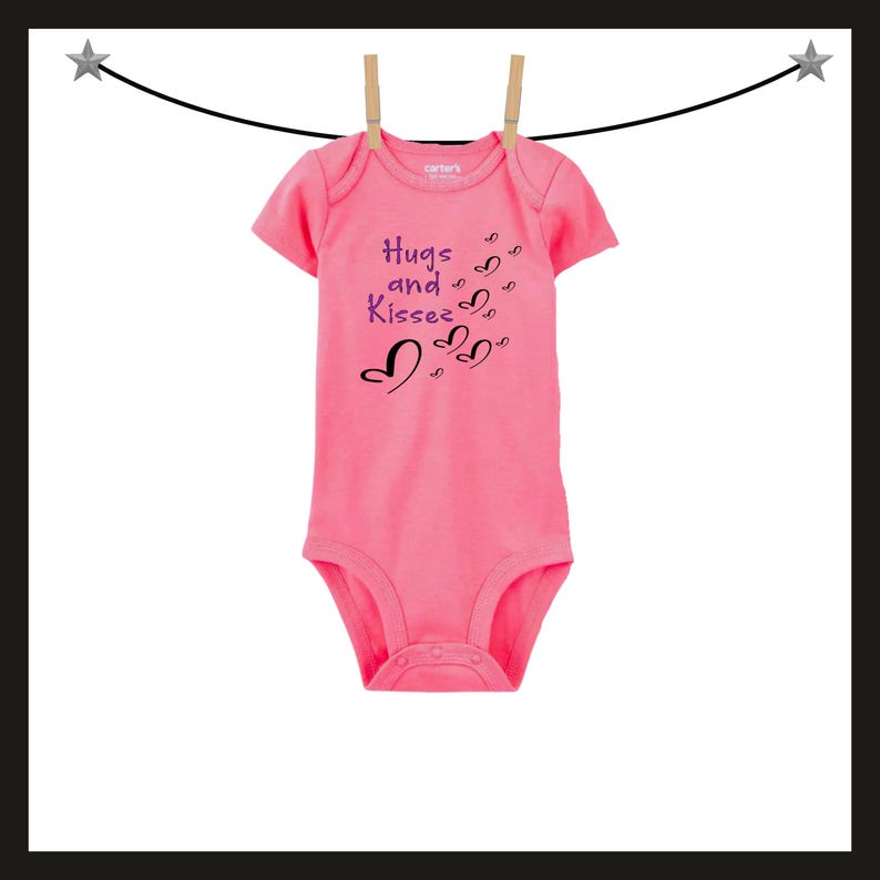 Pode incluir: Um body rosa para beb&eacute; com as palavras "Hugs and Kisses" e desenhos de cora&ccedil;&otilde;es estampados na frente. O body est&aacute; pendurado num varal com molas de madeira. O body &eacute; de manga curta e tem fecho de press&atilde;o.