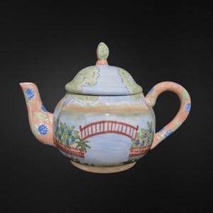 May include: A ceramic teapot with a lid, featuring a painted landscape scene. The design includes a red bridge over water, blue flowers, and a peach-colored handle and spout. The lid has a green leaf-shaped handle.