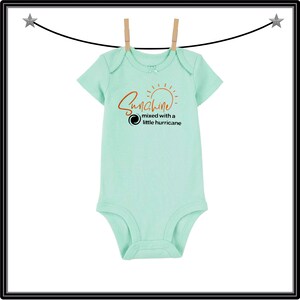 Może przedstawiać: Jasnozielony pajacyk dla niemowląt z napisem "Sunshine mixed with a little hurricane" w kolorze pomarańczowym i czarnym. Pajacyk wisi na sznurku z drewnianymi klamerkami.