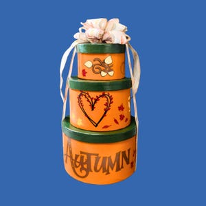 May include: A stack of three round orange boxes with dark green lids, decorated with painted autumn motifs including acorns, leaves, and a heart. The bottom box features the word AUTUMN in dark brown lettering, topped with a decorative fabric bow.