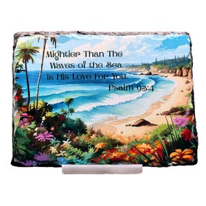 May include: A rectangular decorative stone with a beach scene, palm trees, and colorful flowers. The text reads "Mightier Than The Waves of the Sea is His Love For You. Psalm 93:4". The stone is supported by a silver stand.