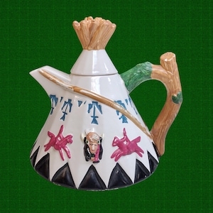 May include: A white ceramic teapot shaped like a teepee, featuring Native American-themed designs. The teapot has a brown handle, a spout, and a lid with a straw-like top. Decorative elements include pink figures and a Native American face.