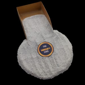 May include: A pile of fine, light gray sand is shown, with a cardboard scoop pouring the sand. A circular sticker with the words "FINE SAND" is visible. The background is black.