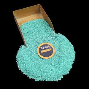 Könnte beinhalten: Eine Schaufel mit teal-grünem Sand mit einem Etikett mit der Aufschrift "0,5-1 MM". Der Sand befindet sich in einer goldenen Metallschaufel.