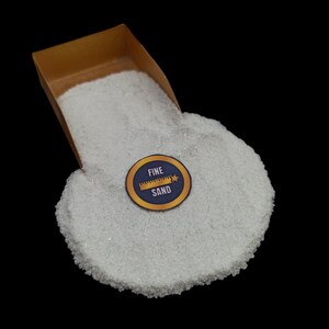 May include: A small, gold-colored scoop filled with fine white sand. A blue and gold sticker with the words "FINE SAND" and a ruler icon is on the sand.