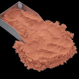 Puede incluir: Una pala de metal llena de un montón de polvo suelto de color marrón rojizo. El polvo se derrama de la pala sobre una superficie negra.