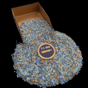 May include: A close-up of a pile of colorful, iridescent glass pieces, ranging in size from 0.5 to 1 mm. The glass pieces are spilling out of a gold-colored container. The label on the container reads "0.5-1MM".