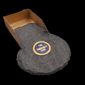 May include: A gold-colored scoop filled with fine black sand. A black and gold sticker with the text "FINE SAND" and a ruler icon is on the sand.