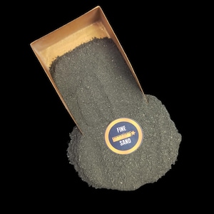 Puede incluir: Una pala de cobre llena de arena verde fina, que se derrama sobre un fondo negro. Una pegatina en la arena dice "ARENA FINA" con una regla y un gráfico de estrella.