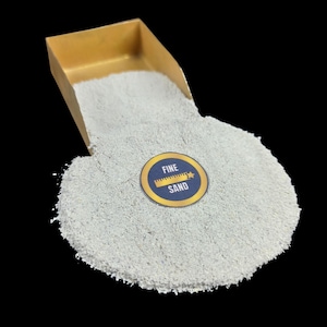 Puede incluir: Se muestra una pila de arena fina, de color gris claro, con una pequeña pala dorada vertiendo la arena. Una pegatina circular con las palabras "FINE SAND" está sobre la arena. El fondo es negro.