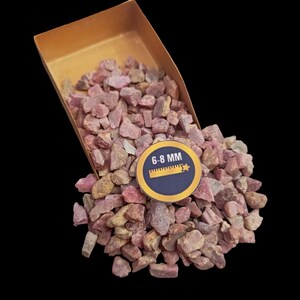 Op de afbeelding: Een close-up van een stapel roze robijn edelsteen chips, variërend in grootte van 6-8 mm. De chips zitten in een bruine metalen schep, die uitloopt op een zwarte achtergrond.