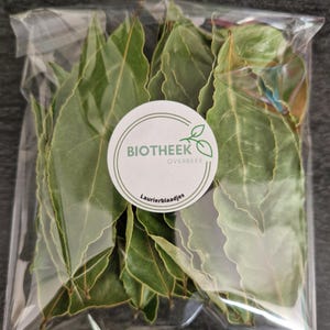 Op de afbeelding: Een doorzichtige plastic zak gevuld met gedroogde laurierblaadjes. De zak heeft een wit label met de tekst "Biotheek Overbeek" en "Laurierblaadjes".