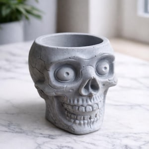 May include: A gray concrete skull-shaped planter with a wide open mouth and eye sockets. The planter has a textured surface and is designed to hold small plants or succulents. The skull is a decorative item.